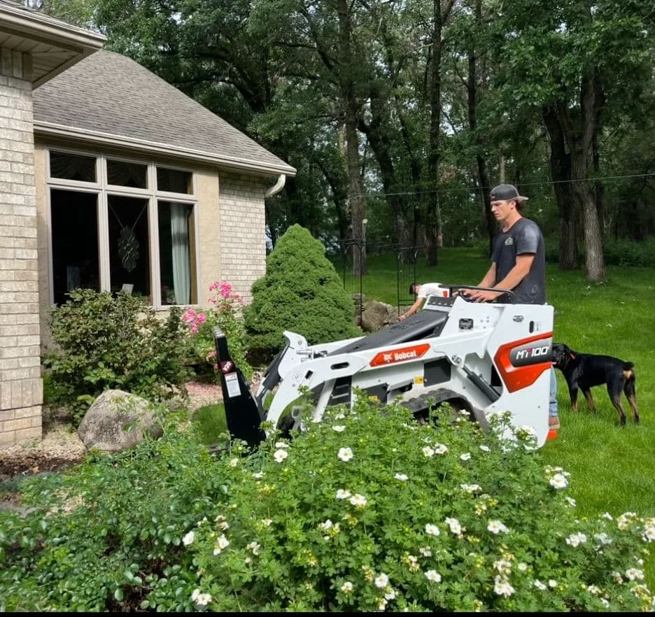 Person operating a skid steer loader for landscaping near a home, with a dog nearby.