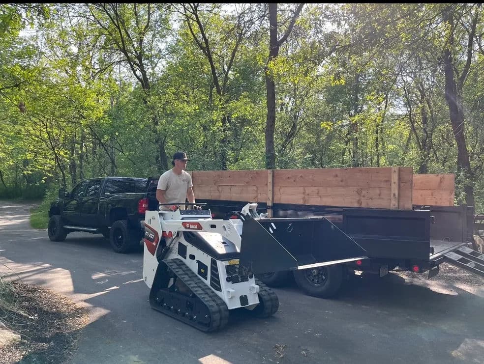 Man operating a compact track loader near a pickup truck and trailer in a wooded area.