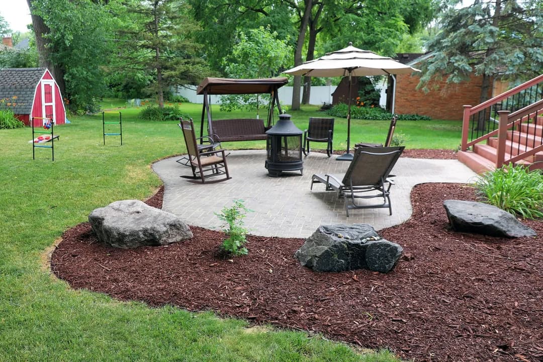 Outdoor patio with seating, fire pit, and umbrella, surrounded by landscaped greenery.