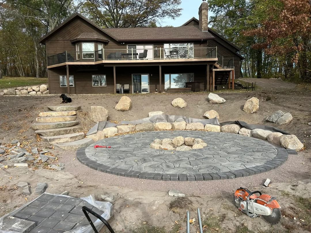 Home renovation with circular stone patio, landscaped yard, and autumn foliage in background.