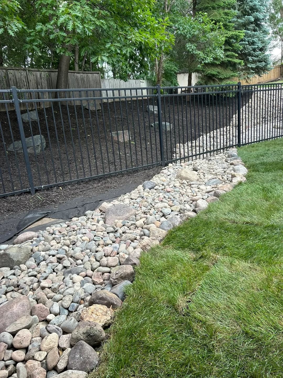 Rock-lined landscape border with green grass and a black metal fence in a shaded yard.
