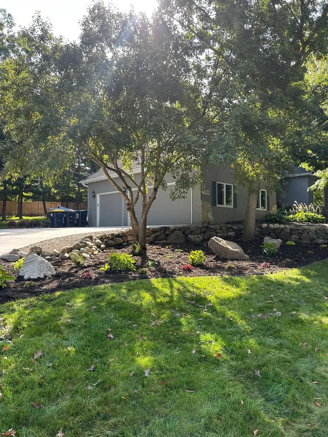 Modern gray house surrounded by lush landscaping and trees, with a stone border and sunlight.
