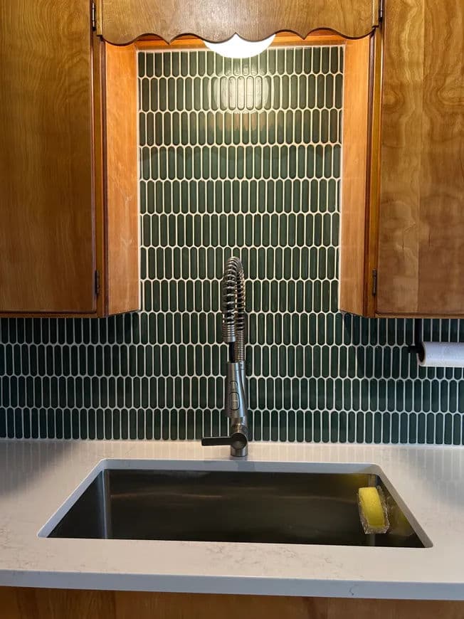 Modern kitchen sink with black basin and green tile backsplash under warm lighting.