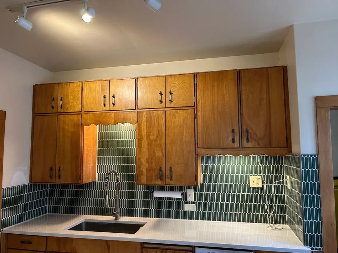 Modern kitchen with wooden cabinets, teal backsplash, and sleek sink. Bright lighting enhances design.